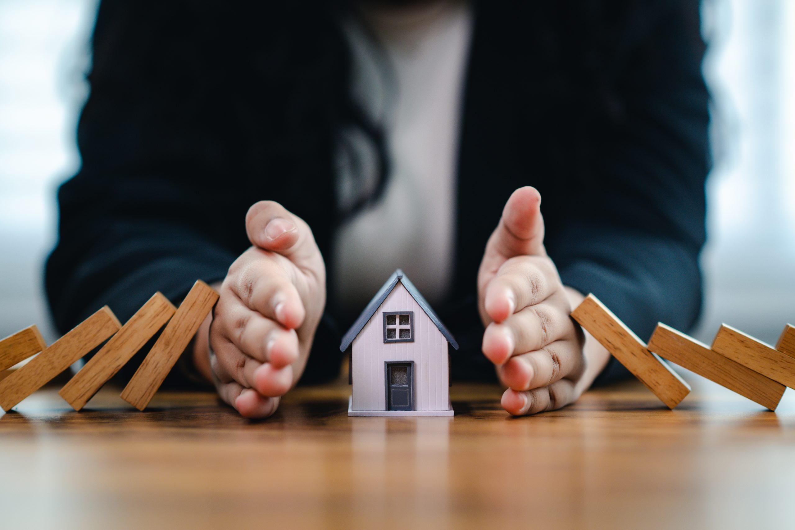 une maison en miniature menacé par une pile de bois qui tombe de chaque coté mais dont les mains d'un monsieur vient protéger